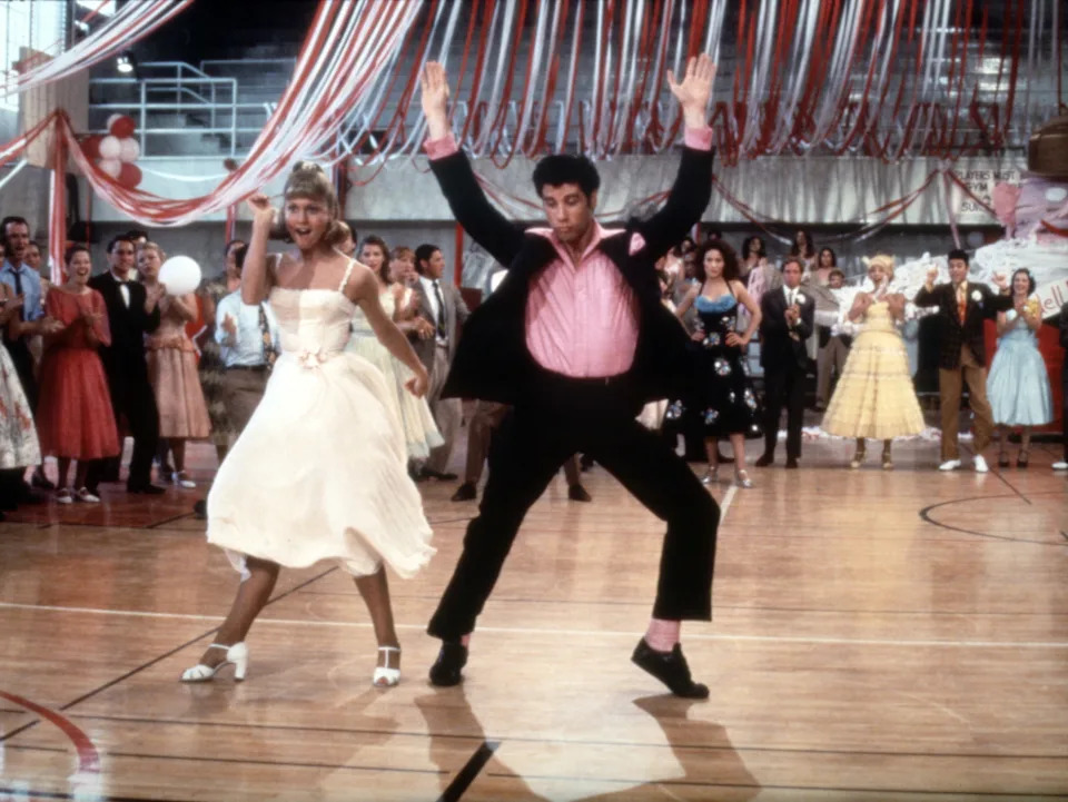 Two people in 1950s-style attire energetically dancing in a school gym decorated with streamers, surrounded by others watching