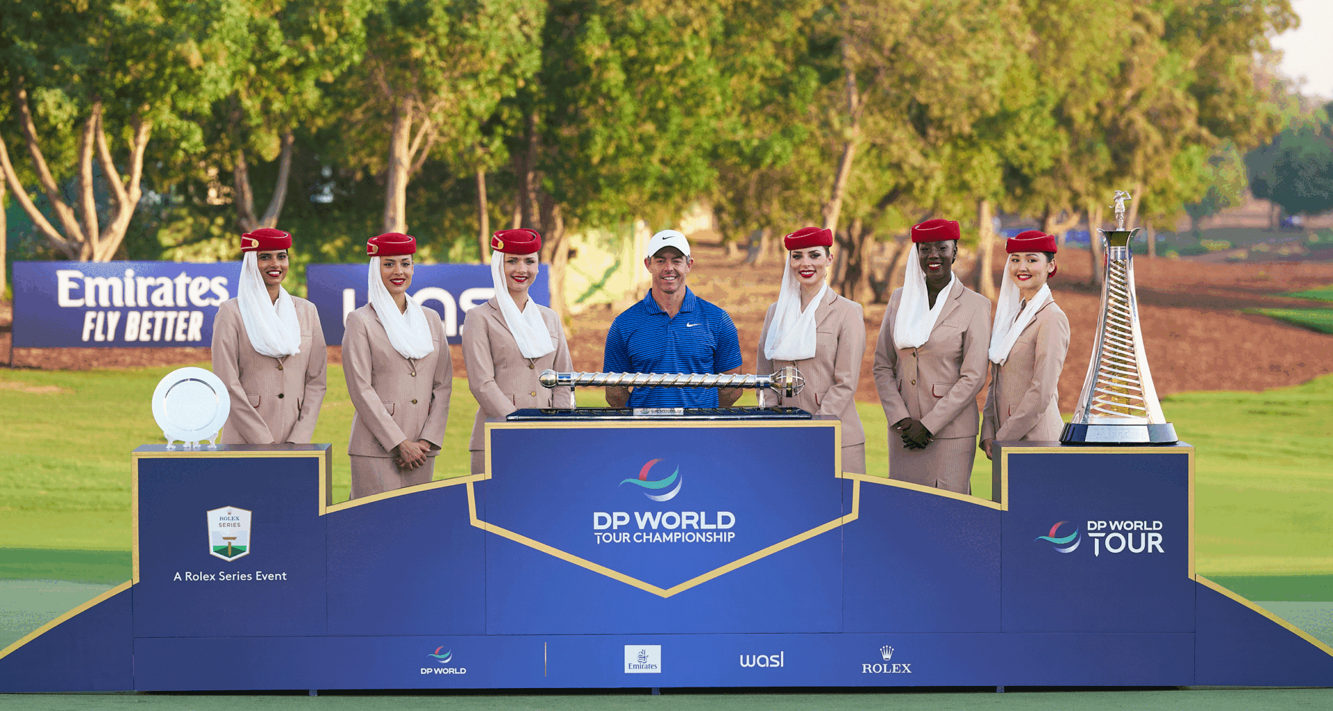 Rory McIlroy stands behind the DP World Tour Championship and the Race To Dubai trophies with six Fly Emirates flight attendants alongside him in 2024