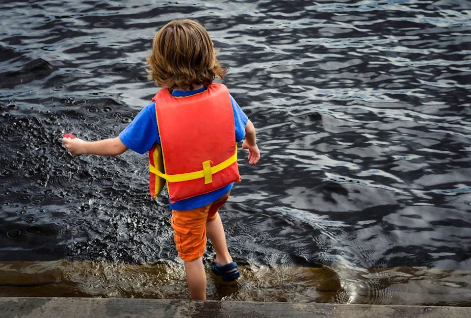Child wearing a life jacket playing by a lake, stepping into the water from a wooden dock