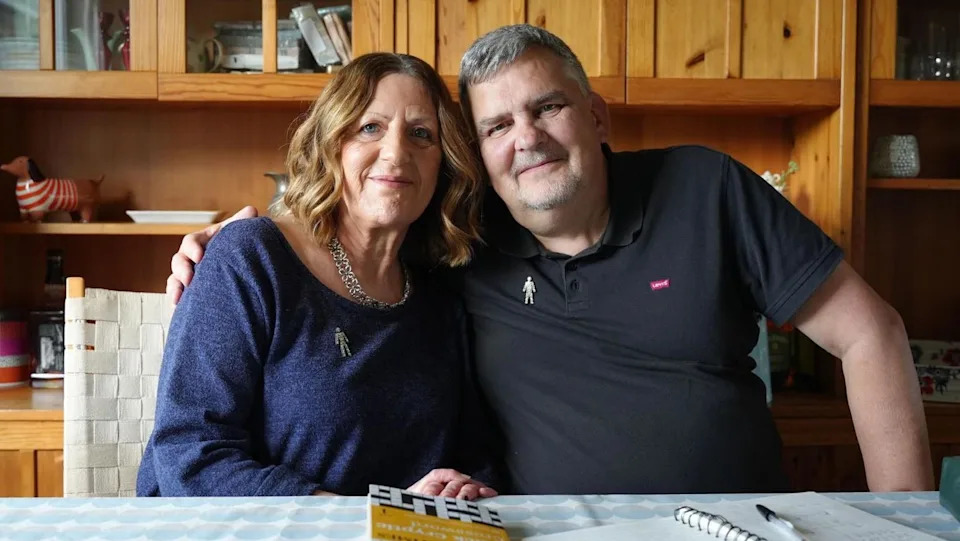 Man and woman both dressed in blue sitting at a dining table, looking at the camera. There is a book of crosswords and pen and paper on the table. Behind them is a wooden cabinet. 