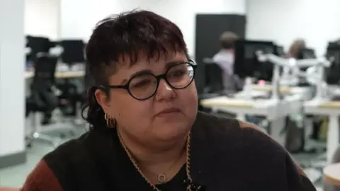 A female presenting person with dark rimmed glasses, a nose piercing, and hooped earings, and shortish dark hair - they are sitting being interviewed in a computer room 