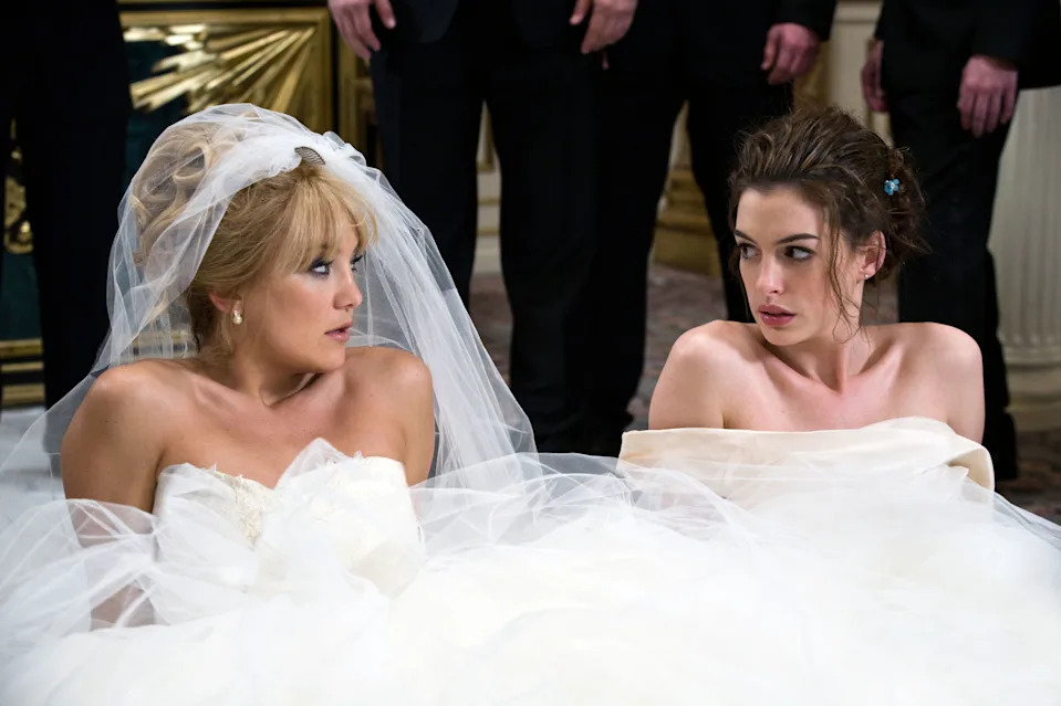 Two women in strapless wedding dresses are sitting on the floor, looking at each other with surprise