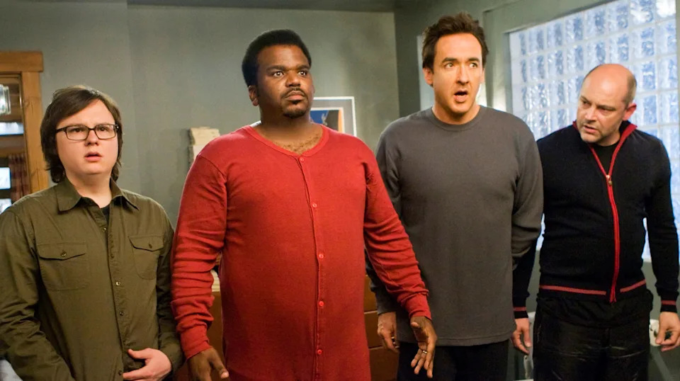 Four men stand indoors, looking surprised. One wears a red sweater, while the others are dressed in casual, dark clothing