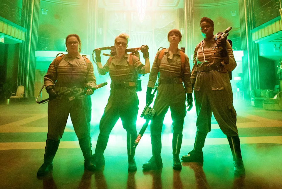Four people in uniforms stand confidently with gear in front of glowing light in a grand hall, resembling a film scene with a heroic vibe