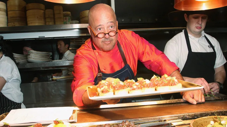 Andrew Zimmern wearing an orange chef jacket, glasses, and apron holds a tray of food.
