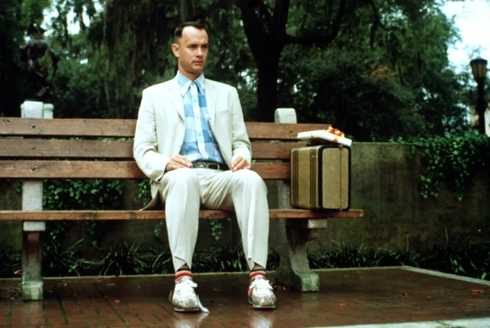 A man in a suit with a plaid shirt sits on a park bench beside a suitcase and box of chocolates