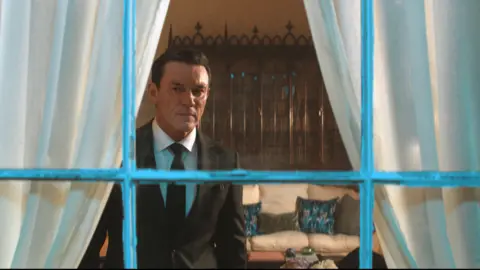 WorldProductions/PA Luke Evans is standing in a living room, wearing a suit and tie, while looking out of a window. Behind him is a sofa and cushions.