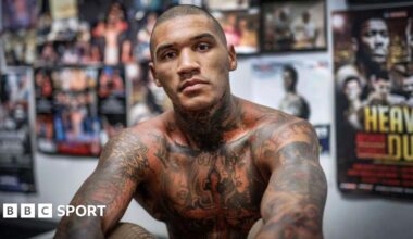 Conor Benn is shirtless and sitting in front a wall of posters