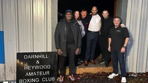 Darnhill and Heywood Amateur Boxing Club Five men and a woman who are on the committee of the community interest group raising money for the project stand in the entrance of the building with a black sign which reads: Darnhill and Heywood Amateur Boxing Club. They are all smiling.