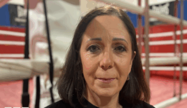 Kerry Norfolk was stood in the middle of the image looking towards the camera. She had shoulder length dark brown hair and was wearing a black jumper. She was stood in front of a boxing ring which had red floors and walls.