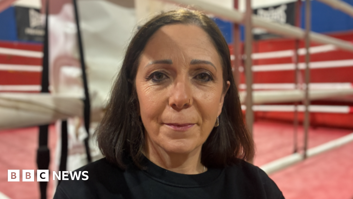 Kerry Norfolk was stood in the middle of the image looking towards the camera. She had shoulder length dark brown hair and was wearing a black jumper. She was stood in front of a boxing ring which had red floors and walls.