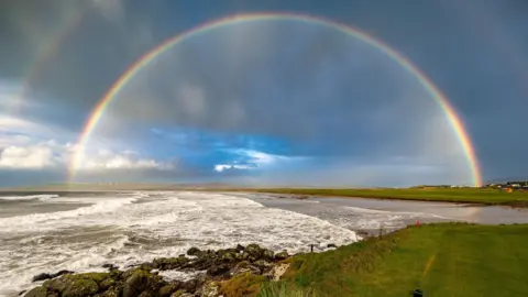 Robert Westerman A sweeping rainbow arcs across a dramatic coastal scene, with waves crashing against dark, moss-covered rocks. The foreground features lush green grass leading to the edge of the shore, contrasting with the stormy sky above. Hints of blue sky peek through the clouds.