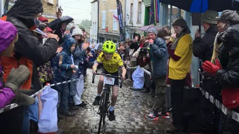 BBC A boy in a yellow T-Shirt and helmet is racing up a narrow path with people standing on either side, cherring him on.