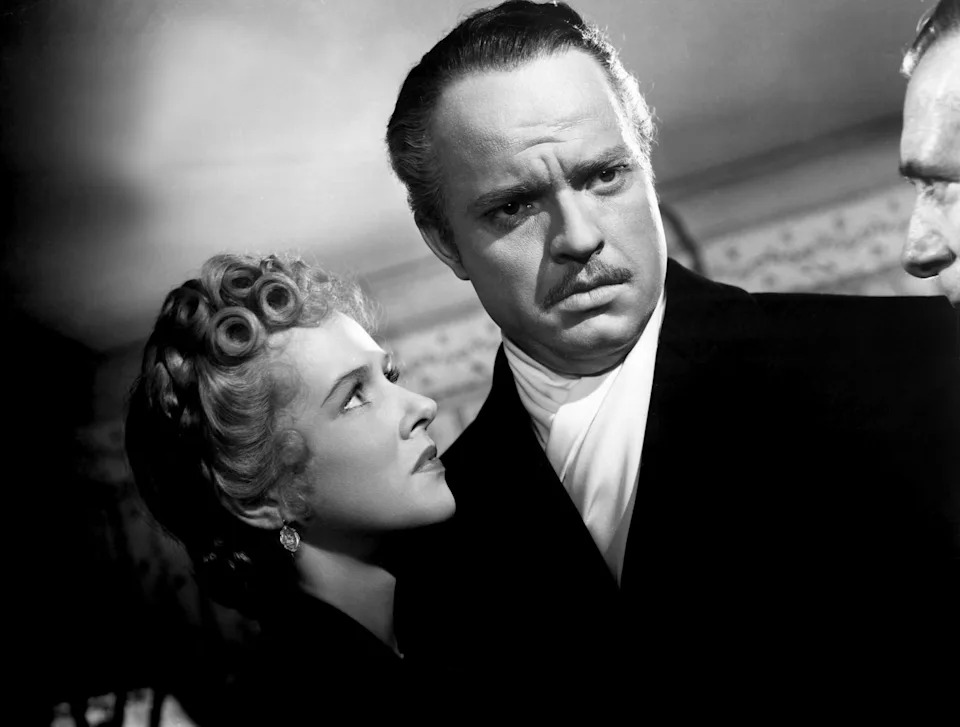 A man in a suit looks serious, standing beside a woman with styled hair, gazing intently at him in a dramatic scene