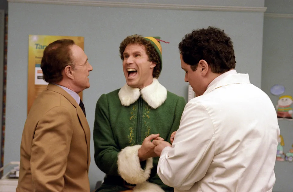 Three people in a playful scene; the central figure wears an elf costume, interacting energetically with the others dressed in a suit and medical attire