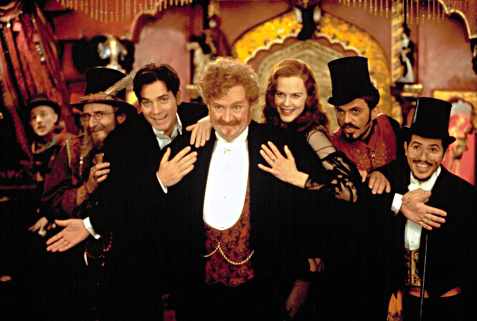 Group dressed in vintage theatrical costumes with top hats and suits, smiling and posing dramatically in front of an ornate backdrop