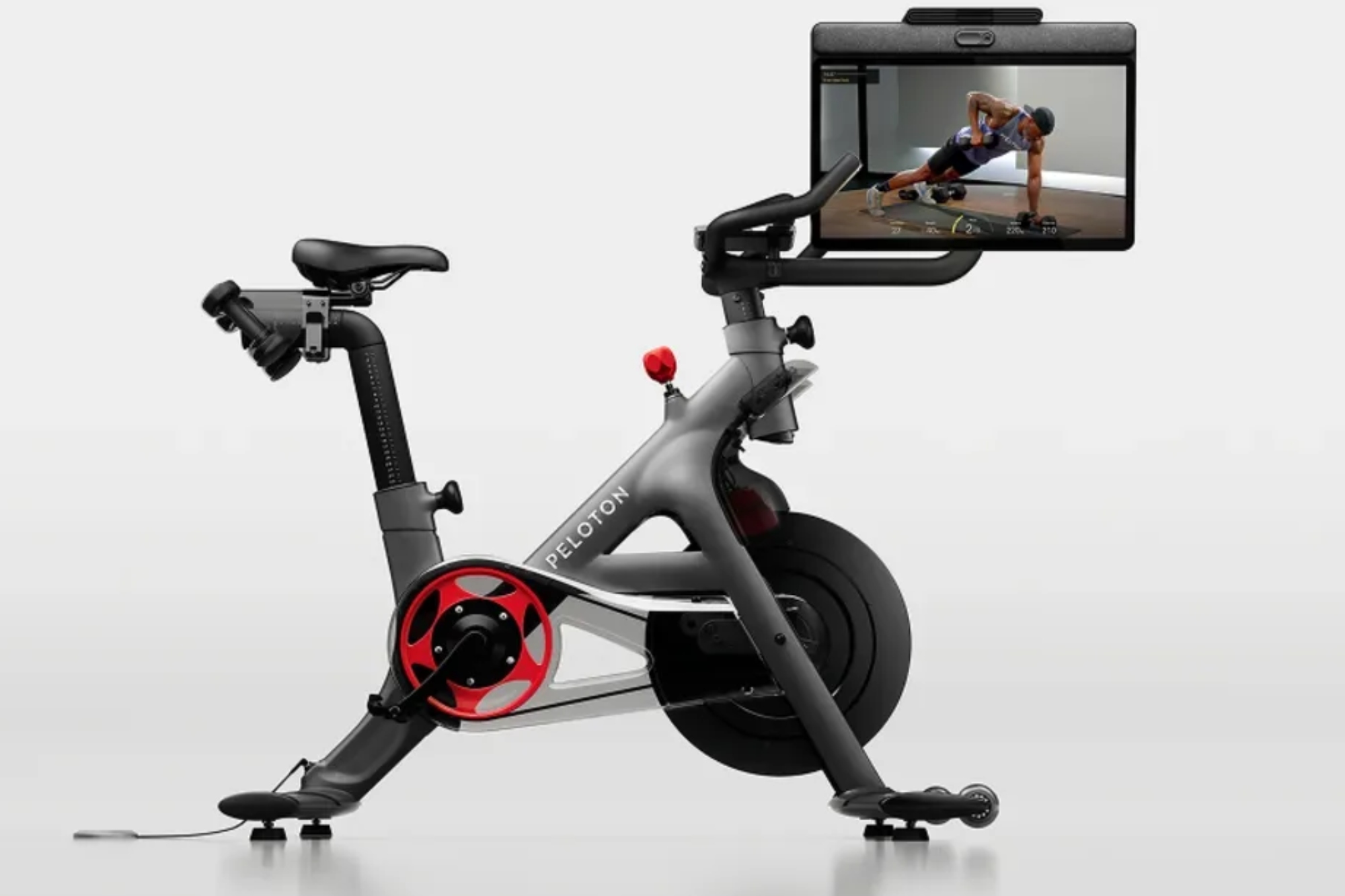 The Peloton Cross Training Bike + in full from the side with the screen showing a white man completing a devel press with dumb bells