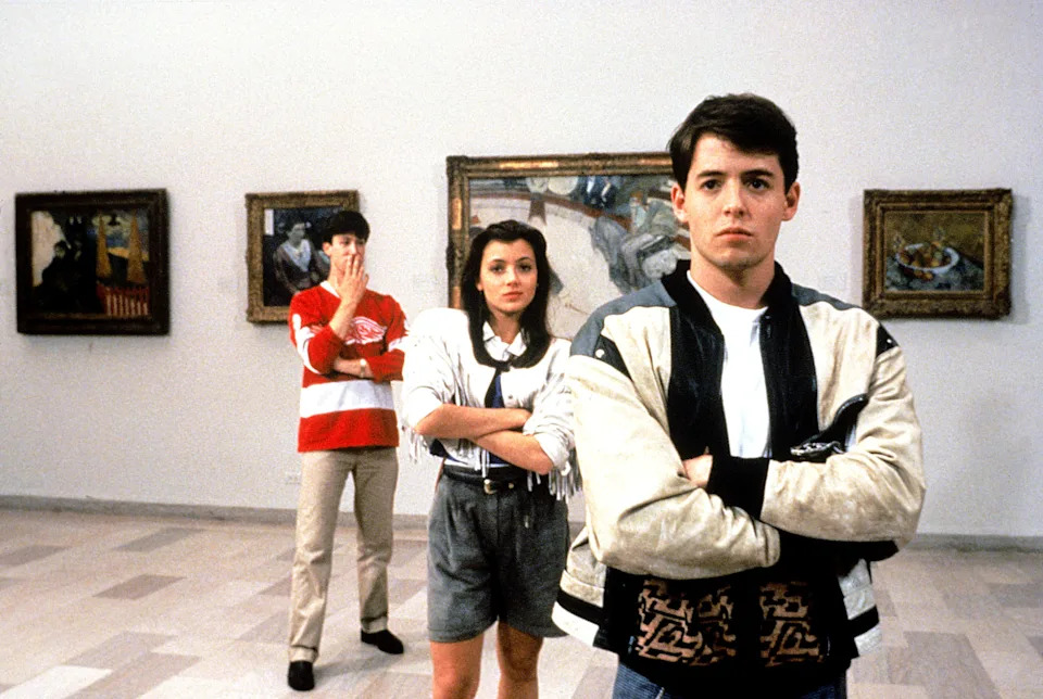 Three young adults are in an art gallery; one wears a varsity jacket, another covers his mouth, and the third stands with arms crossed