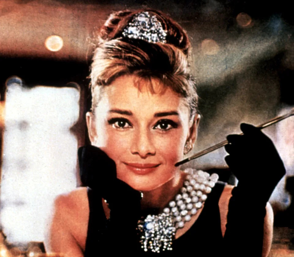 Person in elegant attire with pearl necklace, holding a cigarette holder, posing with a relaxed expression and styled hair