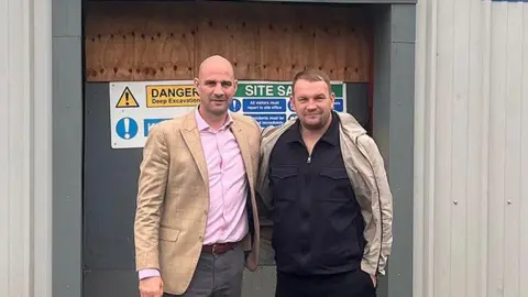 Darnhill & Heywood Amateur Boxing Club Dale Gaucas wearing a pink shirt and beige blazer and grey pants and Danny Randall with brown hair wearing a black shirt and beige coat and black trousers stand arm in arm in front of the building where they are opening the boxing club.  They are smiling.