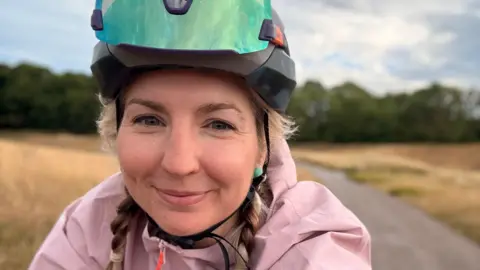 BBC Claire Sharpe wears a cycling helmet and pink jacket has taken a selfie whilst riding her bike through a rural area. She has blonde hair and is smiling. 