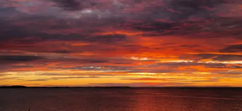 Alan Nairn A vivid sunrise paints the sky in fiery shades of orange, red, and purple, blending into darker clouds above. There is a calm sea below reflecting the sunset. 
