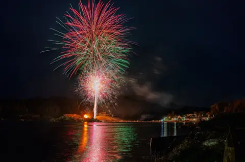 Sam Bilner Brilliant fireworks explode in the night sky, with vibrant red and green streaks radiating outward in a dazzling display. Below, a quiet coastal village glows with warm lights, reflecting softly on the water’s surface.

