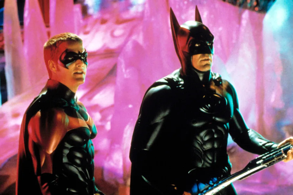 Two actors in superhero costumes resembling Batman and Robin stand in an icy cave setting, appearing focused and ready for action