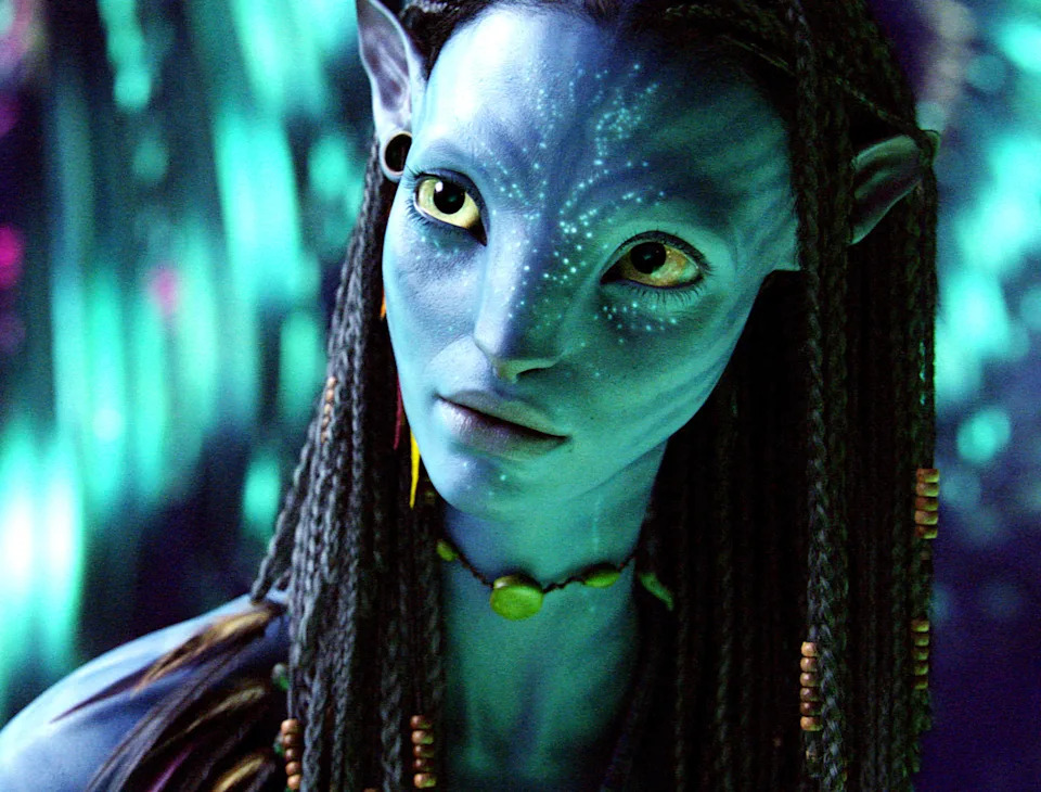An alien character with blue skin, braided hair, and a curious expression appears in a lush environment, possibly from a science fiction film