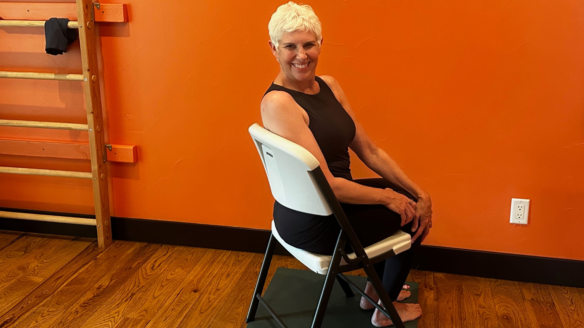 woman in a white chair in a spinal twist position. she's on a wooden floor with an orange wall behind her.