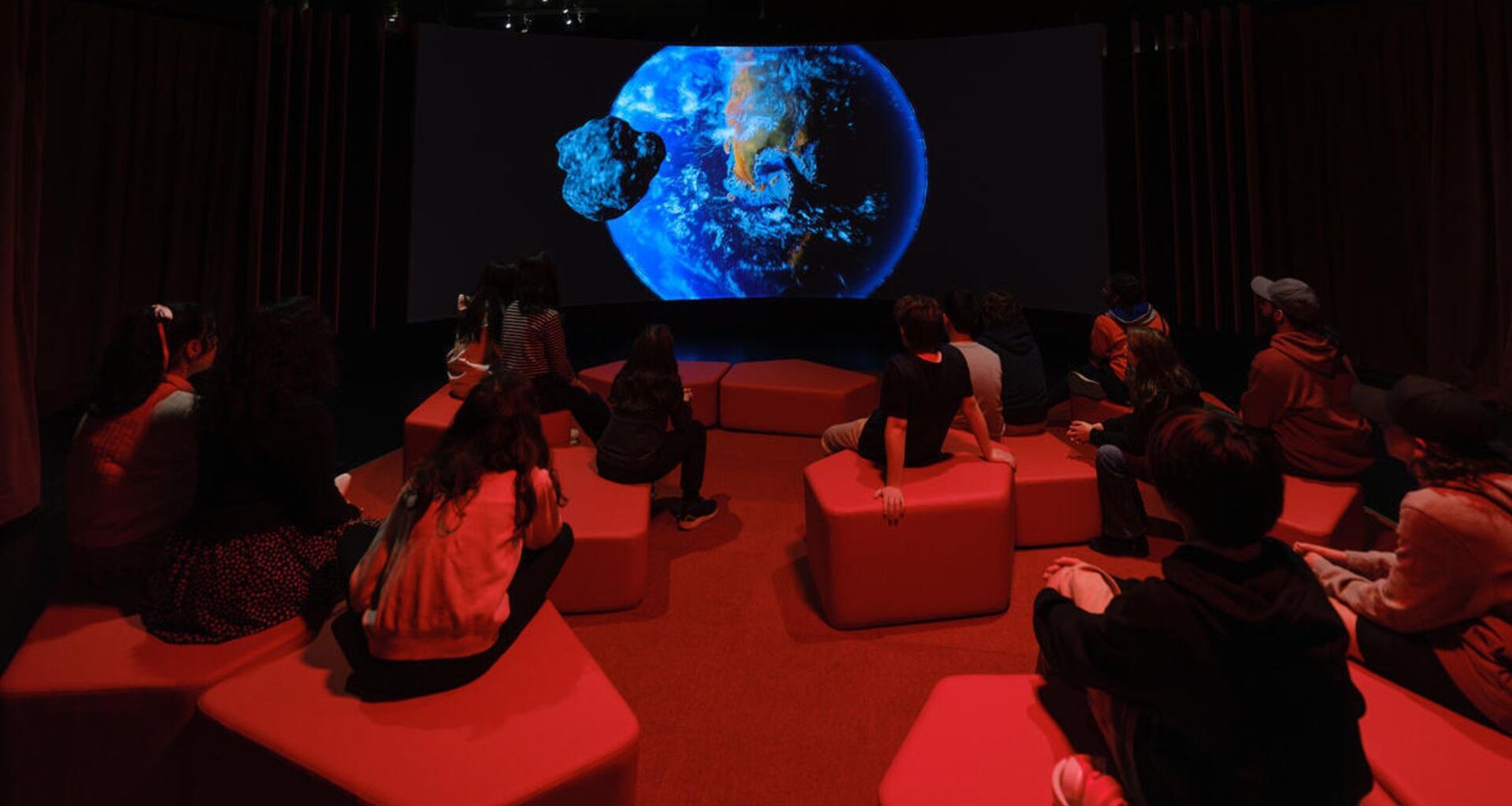 An Earth is projected on a dark screen as children sit on blocks in a dimly red-lit theater