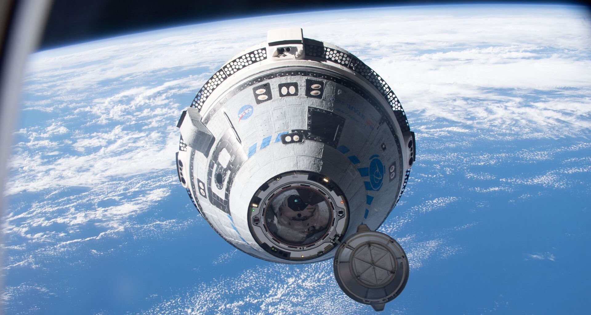 A white and blue Boeing Starliner capsule floats above a blue Earth with the black of space in the background.