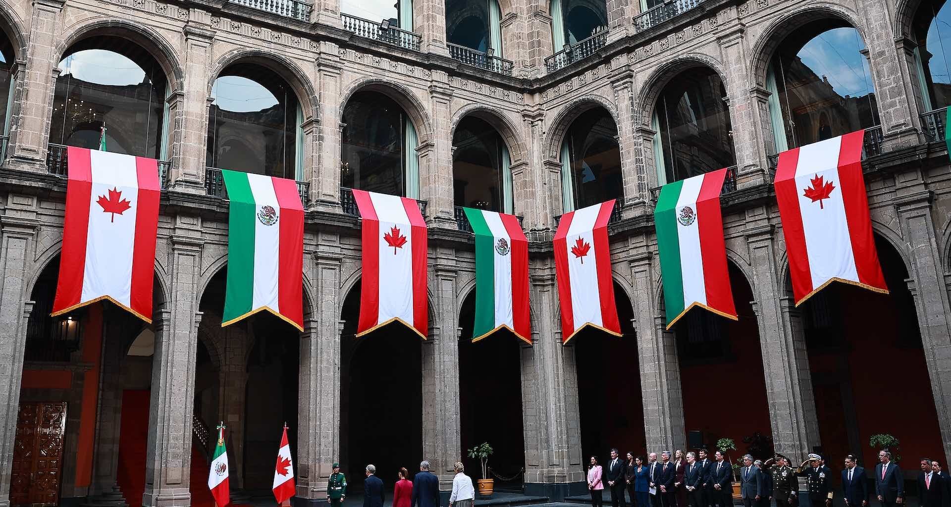A wide shot of the courtyard at the Palacio Nacional in Mexico City, which is draped with large banners bearing the colours and emblems of the Canadian and Mexican flags. Mark Carney and Claudia Sheinbaum are walking toward a red-carpeted entrance, with other dignitaries standing to their right.