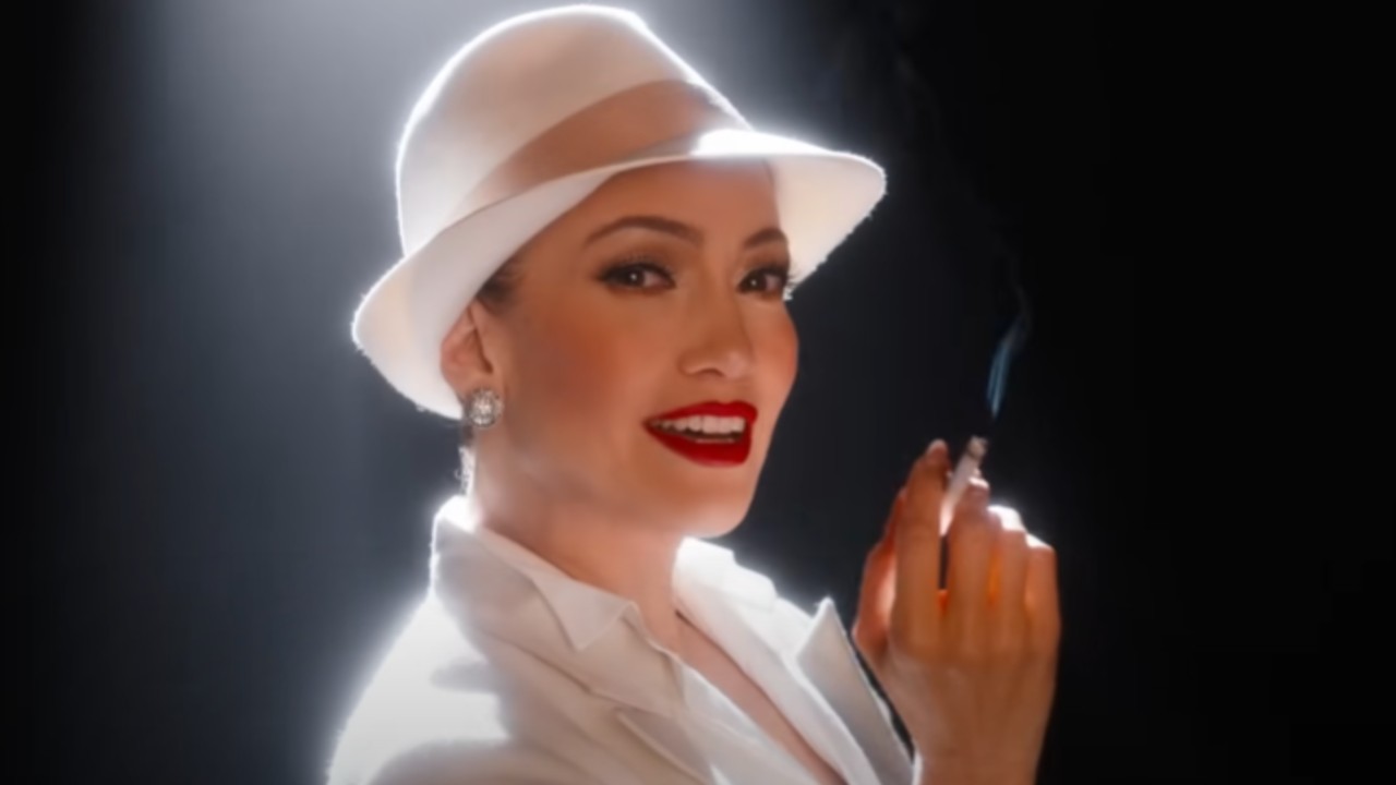 JLo wearing a white fedora in Kiss of the Spider Woman and holding a cigerette in her hand.