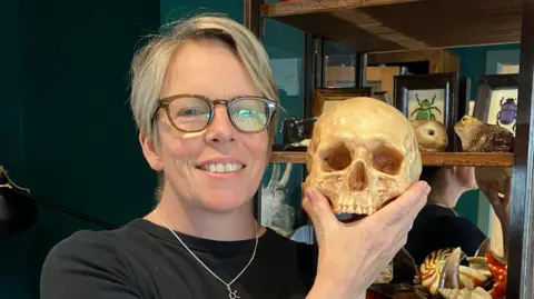 Henry Godfrey-Evans/BBC A woman holding what looks like - but definitely is not - a skull, in the background is a cabinet of beetles, shells and a brain