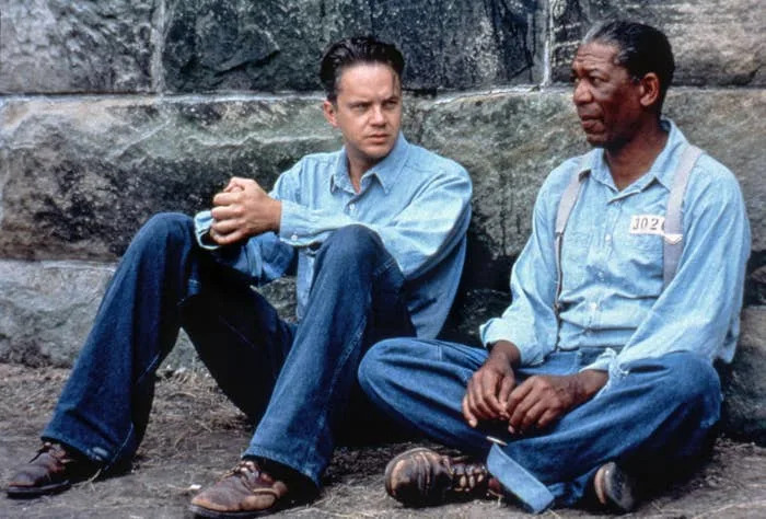 Tim Robbins and Morgan Freeman in uniforms sit on the ground against a stone wall, engaged in a conversation