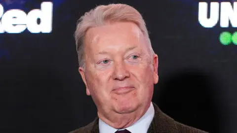 PA Media Promoter Frank Warren looks away at the camera as he slightly smiles. He has short grey hair. He wears a brown suit jacket, white shirt and tie. He is standing in front of a large screen with some brand names on it that cannot be read. 