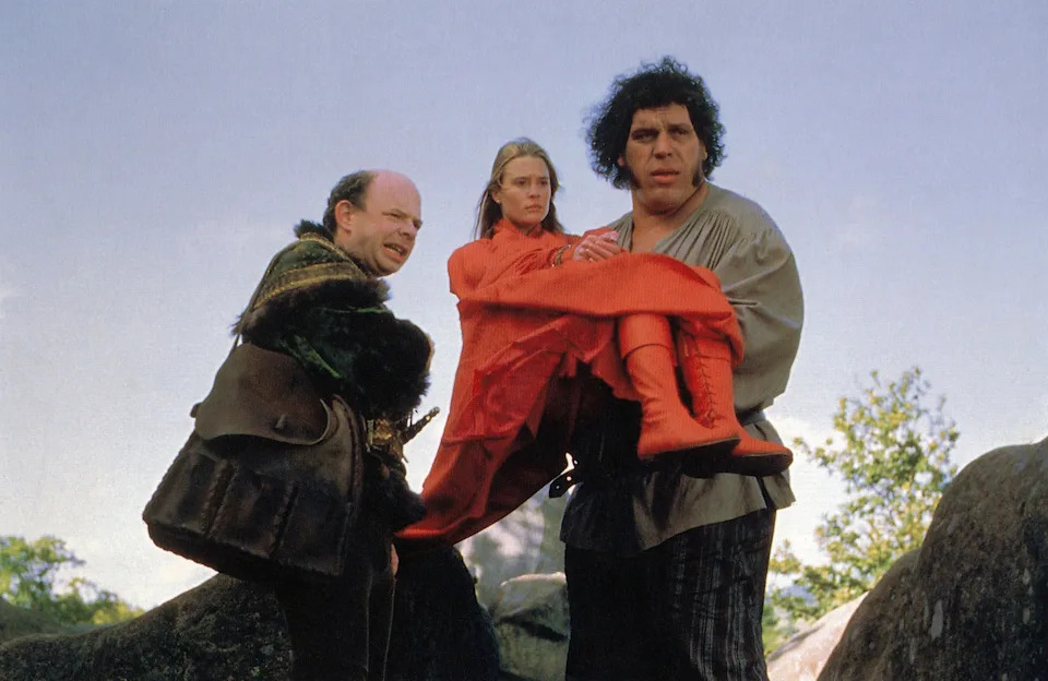 Scene from a fantasy film with a man carrying a woman, another man in medieval-style clothing stands beside them outdoors