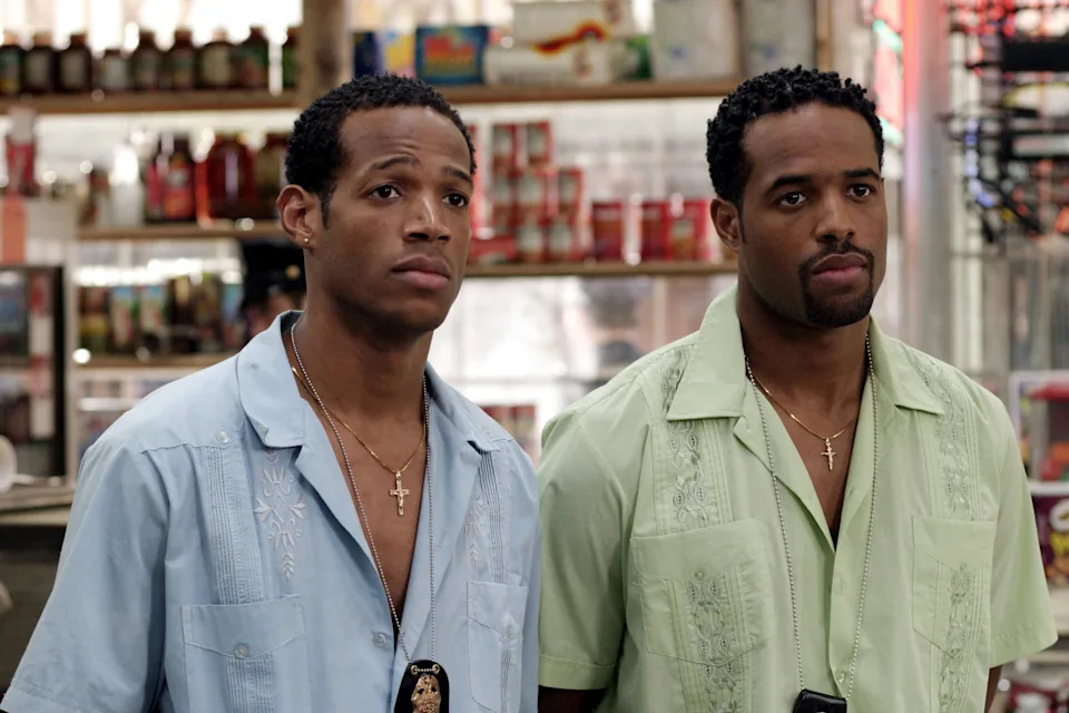 Two people stand in a store wearing embroidered shirts, one in light blue and the other in light green, each with a necklace and serious expressions