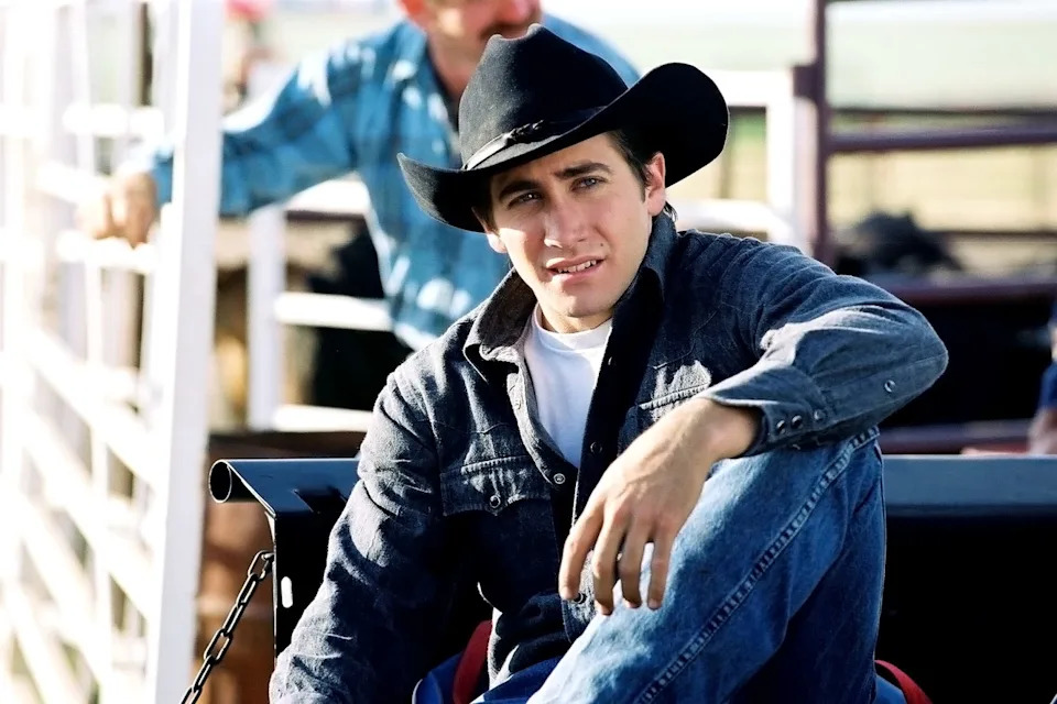 Person in cowboy hat and denim shirt sitting casually on a fence with a thoughtful expression. Background features a blurred figure and fencing
