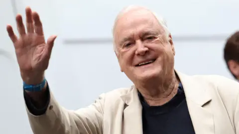 Reuters Actor John Cleese is seen smiling and waving at the camera. He has white hair and is wearning a cream suit jacket, blue watch and black jumper.