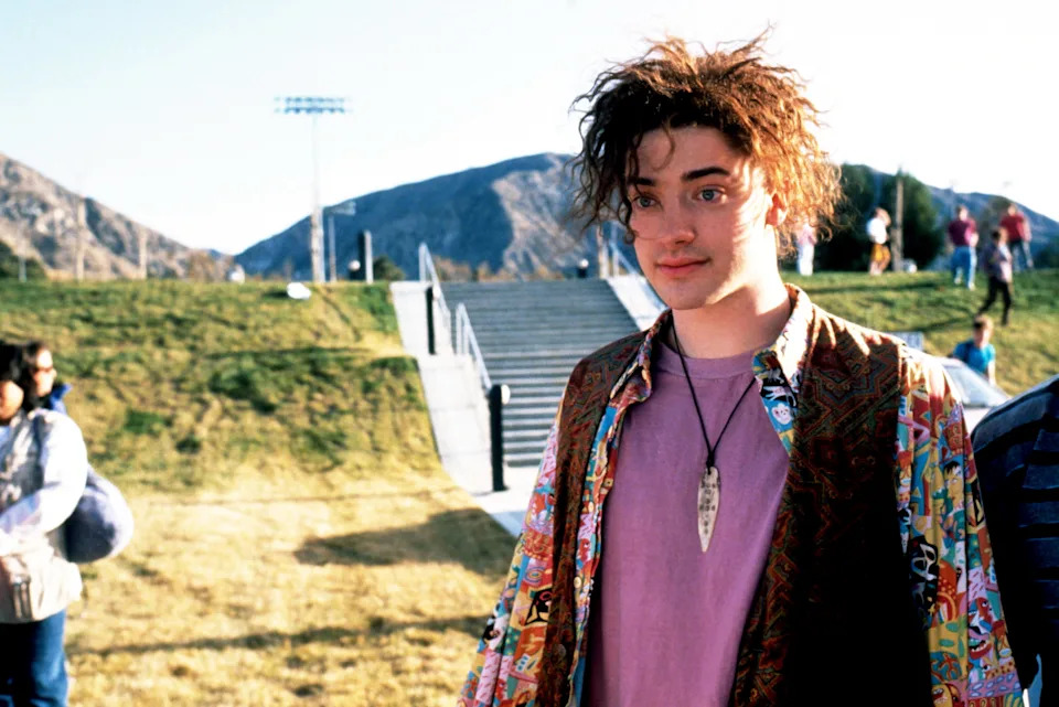A person with wild hair stands outdoors on grass, wearing a patterned shirt over a purple top and a necklace with a pointed pendant. Mountains are in the background
