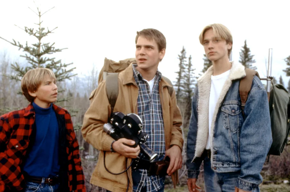 Three young people outdoors in casual clothing with backpacks, one holding a camera. Pine trees are visible in the background