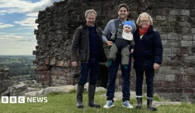A family group standing outdoors in front of the remains of an old stone wall, part of an historic ruin or castle. The people are dressed in outdoor clothing, including jackets and boots, and the man in the centre is carrying a small child. Behind the group there is a scenic view of green fields stretching into the distance under a partly cloudy sky.