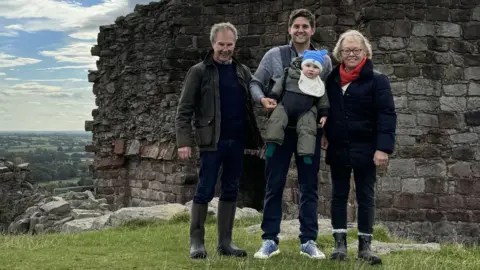 Family photo A family group standing outdoors in front of the remains of an old stone wall, part of an historic ruin or castle. The people are dressed in outdoor clothing, including jackets and boots, and the man in the centre is carrying a small child. Behind the group there is a scenic view of green fields stretching into the distance under a partly cloudy sky. 