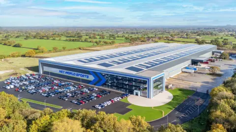 Net World Sports A large modern industrial building with “NET WORLD SPORTS” displayed prominently on the front in bold blue lettering. The structure has a glass-fronted office section on one side and an extensive warehouse area behind. Several rows of parked cars fill the adjacent car park, and a few lorries are positioned near loading bays. The site is surrounded by greenery, with open fields stretching into the distance under a partly cloudy sky.