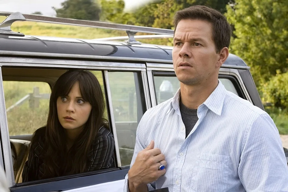Two people outside a vehicle look concerned, one seated inside, wearing a striped shirt, the other outside, in a light button-down shirt