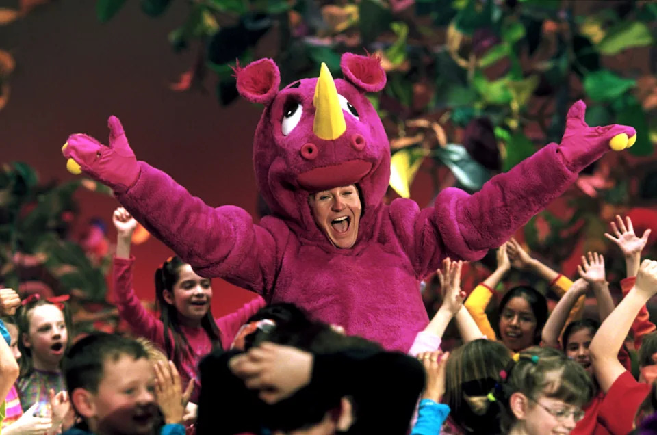 Person in a pink dinosaur costume joyfully surrounded by excited children