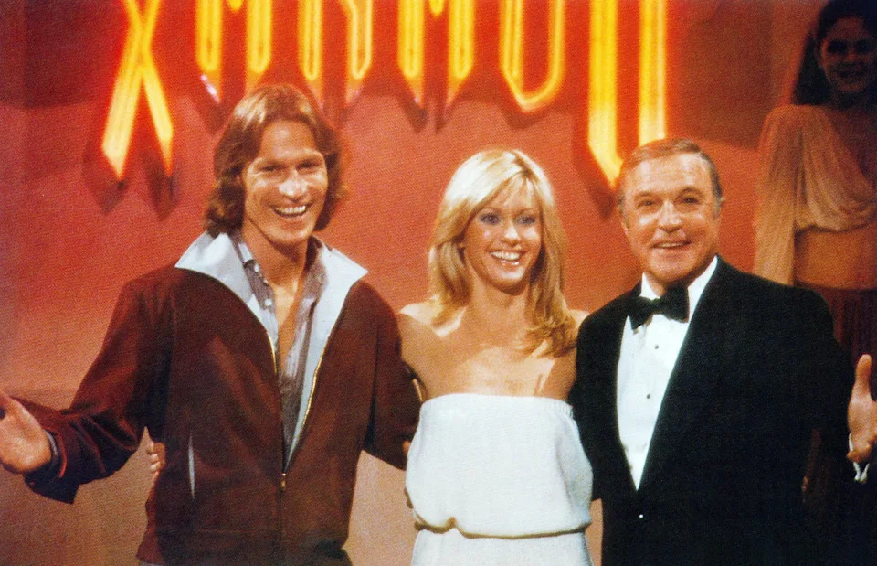 Three people stand together, smiling. The man on the left wears casual attire, the woman in the middle is dressed in an elegant, strapless outfit, and the man on the right wears a tuxedo