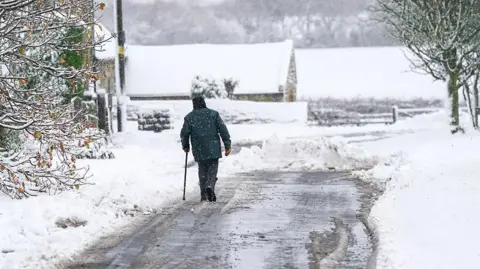 Getty Images Man walks on a road partially cleared of snow, with a covering of snow on trees in the foreground and a barn and fields in the background